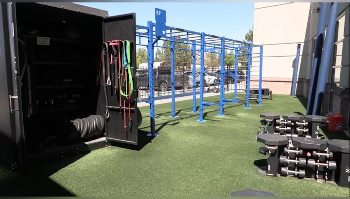 An outdoor gym area features exercise equipment, including free weights, resistance bands, and a bar setup on artificial turf.