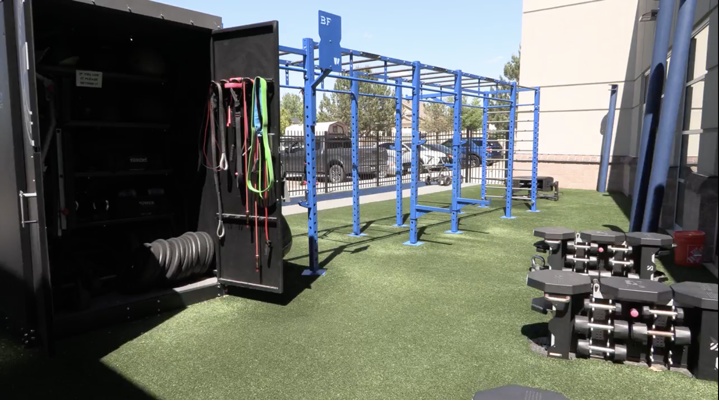 An outdoor gym area features exercise equipment, including free weights, resistance bands, and a bar setup on artificial turf.
