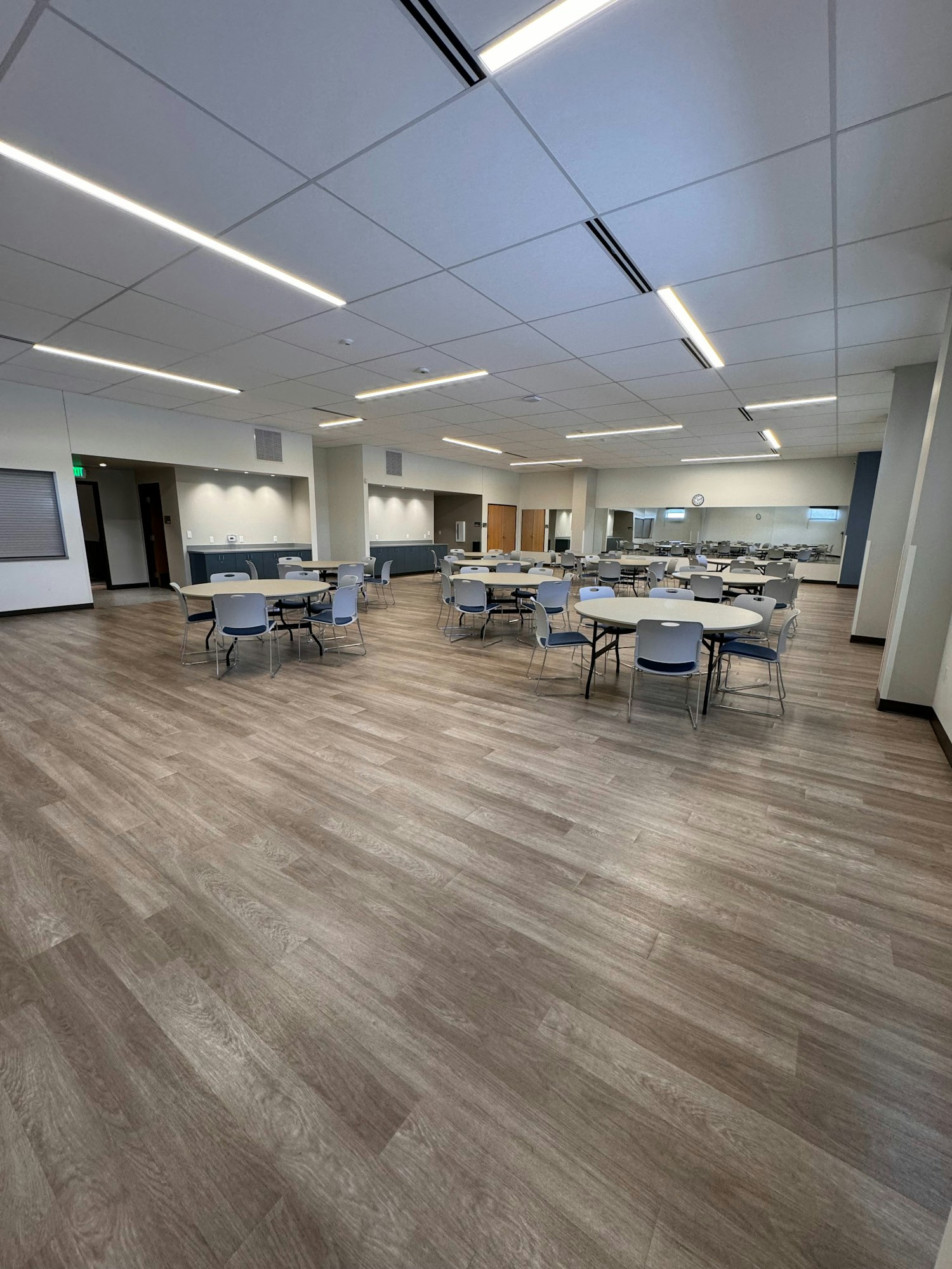 A spacious, empty room with round tables and chairs on a wooden floor, bright overhead lights, and a wall mirror.