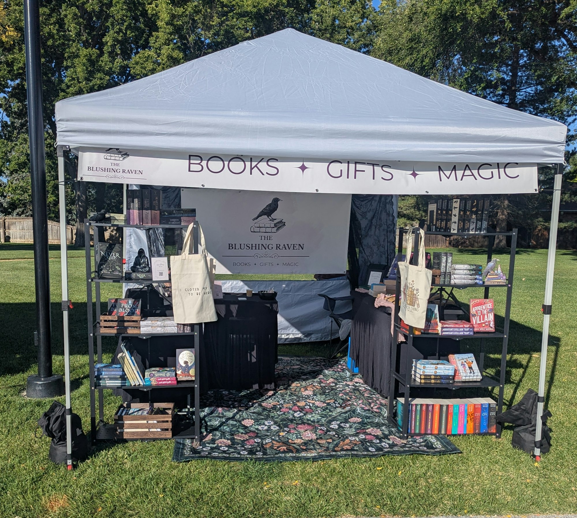 Outdoor book stall with "The Blushing Raven" canopy, showcasing books, gifts, and magic-themed items on shelves.