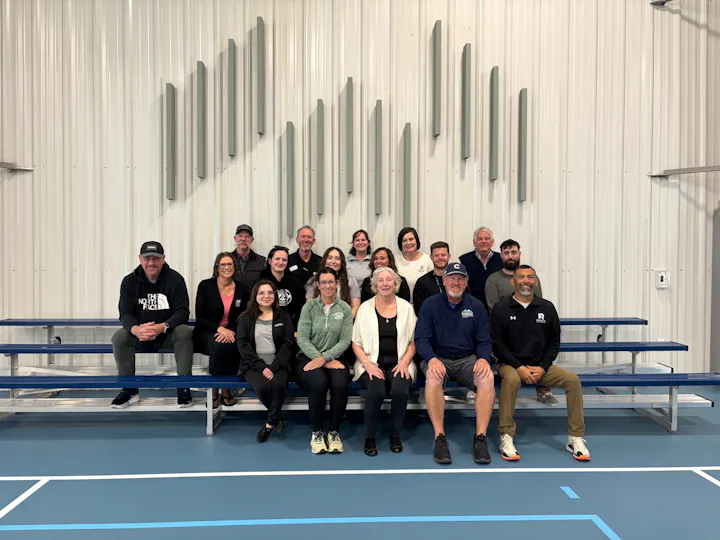 A diverse group of people poses together on bleachers in a gym or sports facility.