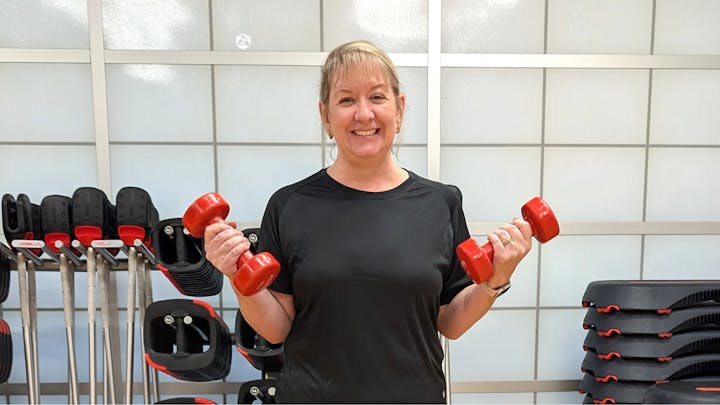 A person in a gym holding red dumbbells, smiling, with fitness equipment in the background.