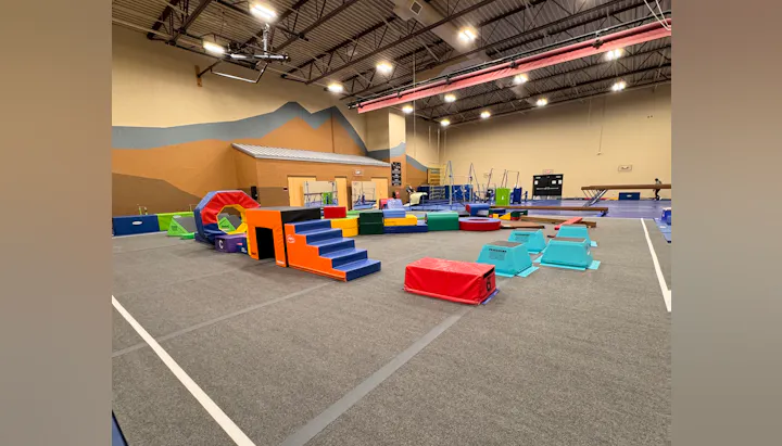 The image shows a colorful gymnastic training area with padded equipment for exercises and obstacles.