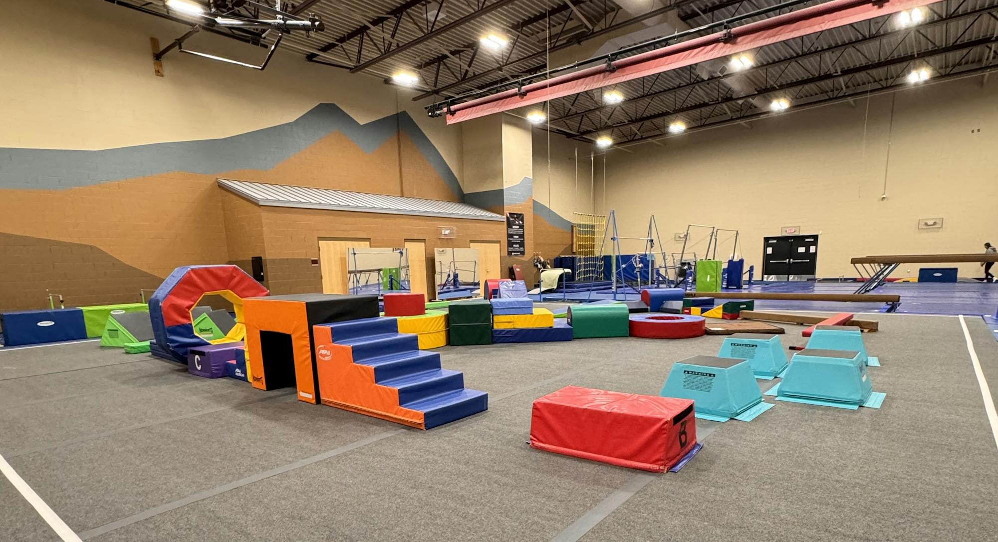 The image shows a colorful gymnastic training area with padded equipment for exercises and obstacles.