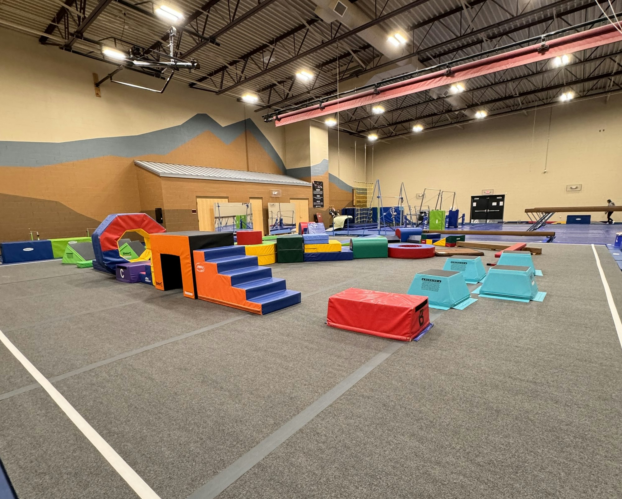 The image shows a colorful gymnastic training area with padded equipment for exercises and obstacles.