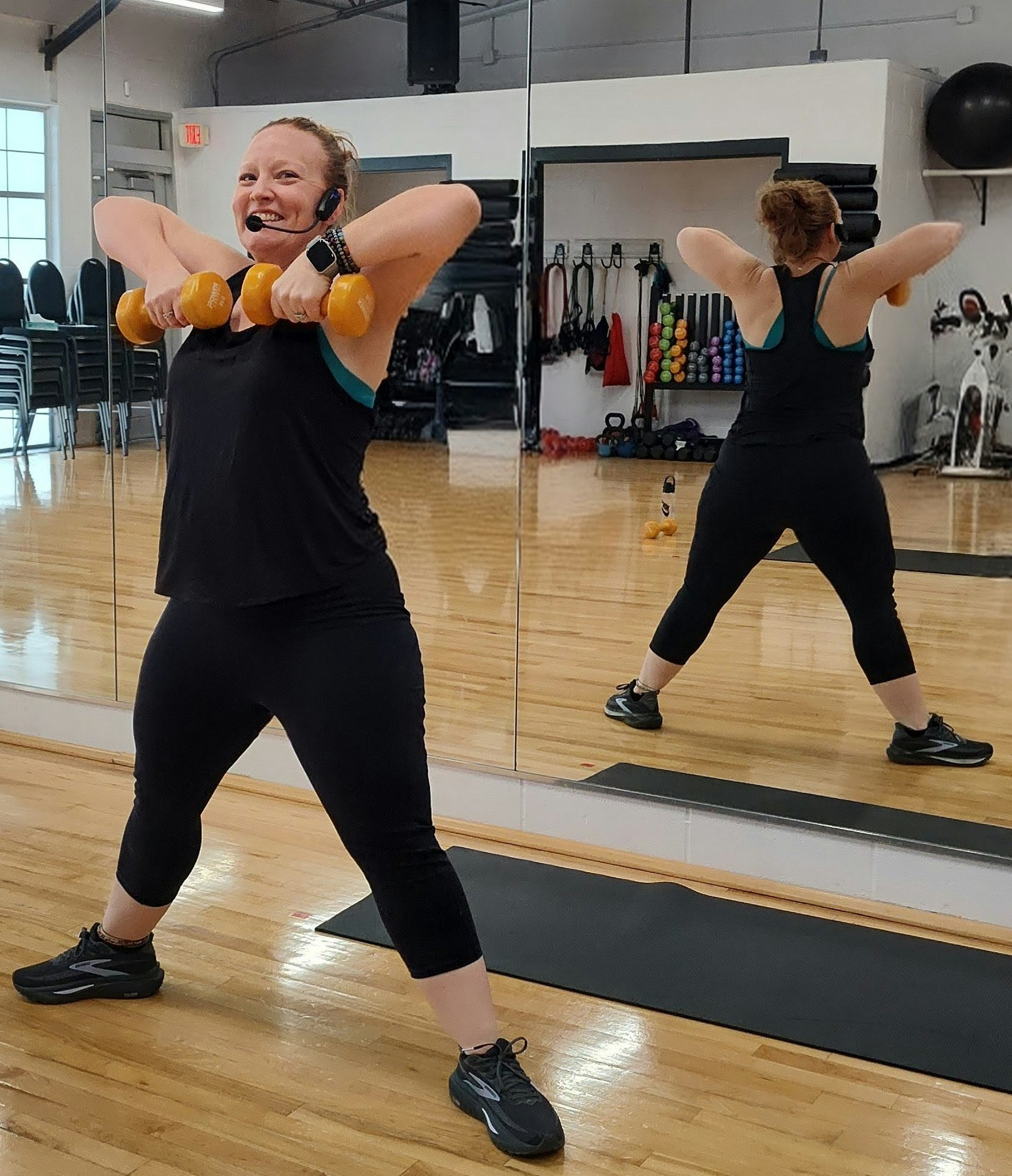 A woman is exercising in a gym, lifting dumbbells and smiling at her reflection in the mirror.