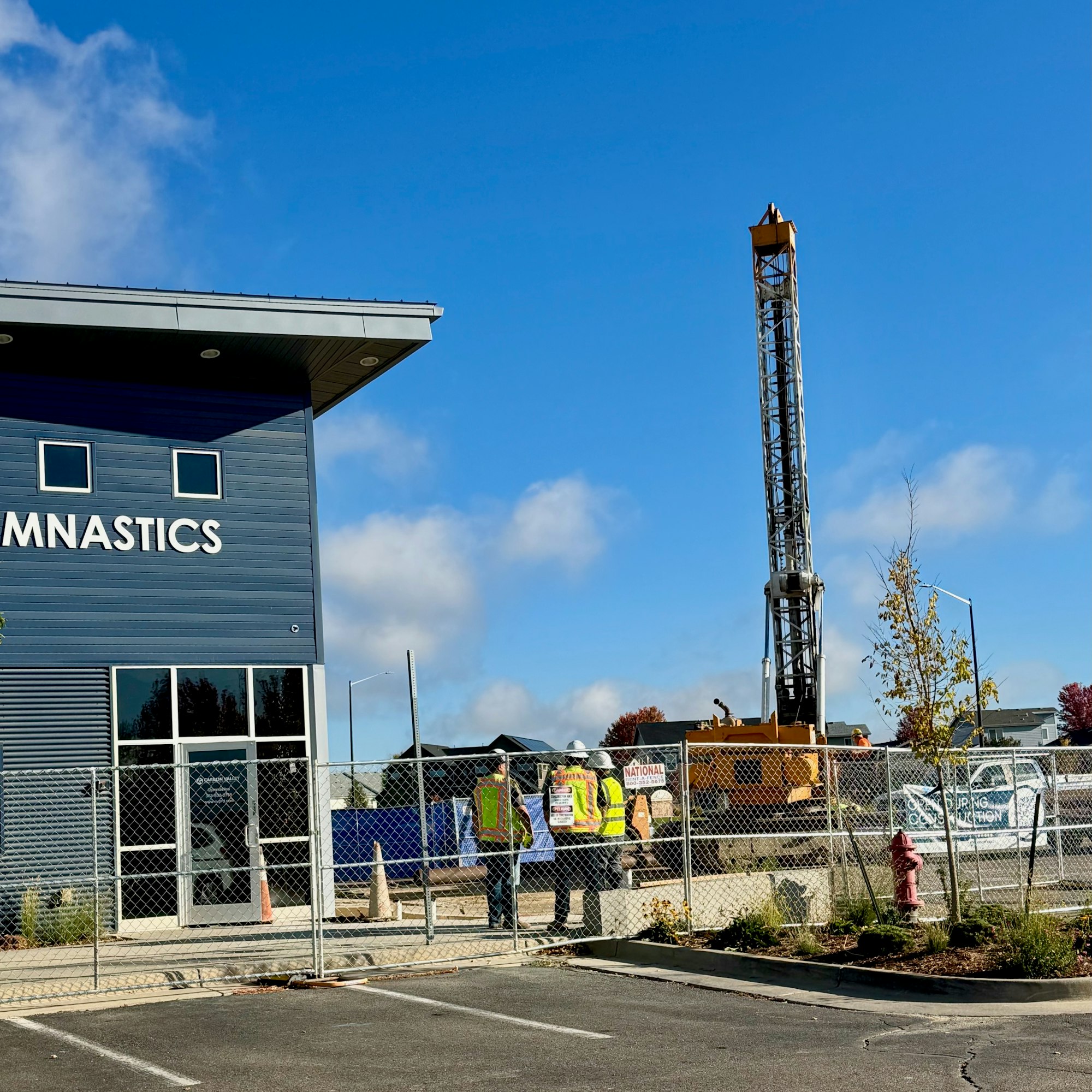 A gymnastics building next to a construction site with workers and machinery, surrounded by a chain-link fence.