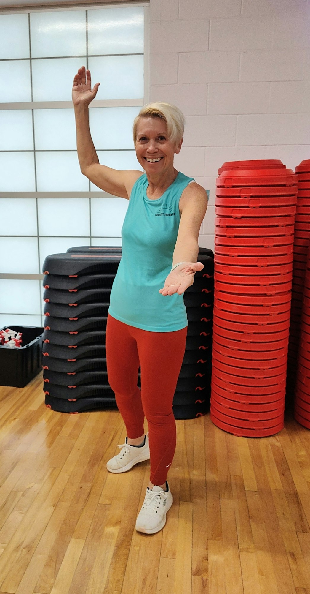 A smiling woman in athletic wear stands with one arm raised and the other extended, surrounded by stacked exercise equipment.