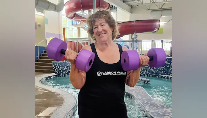 A smiling woman in a swimsuit holds purple dumbbells near a pool, promoting fitness in a recreational setting.