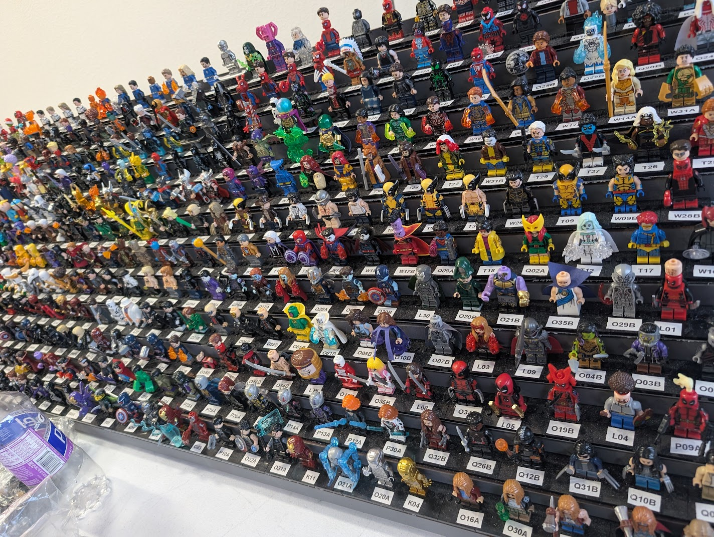 A display of various LEGO minifigures in rows with labeled slots and a water bottle in the corner.