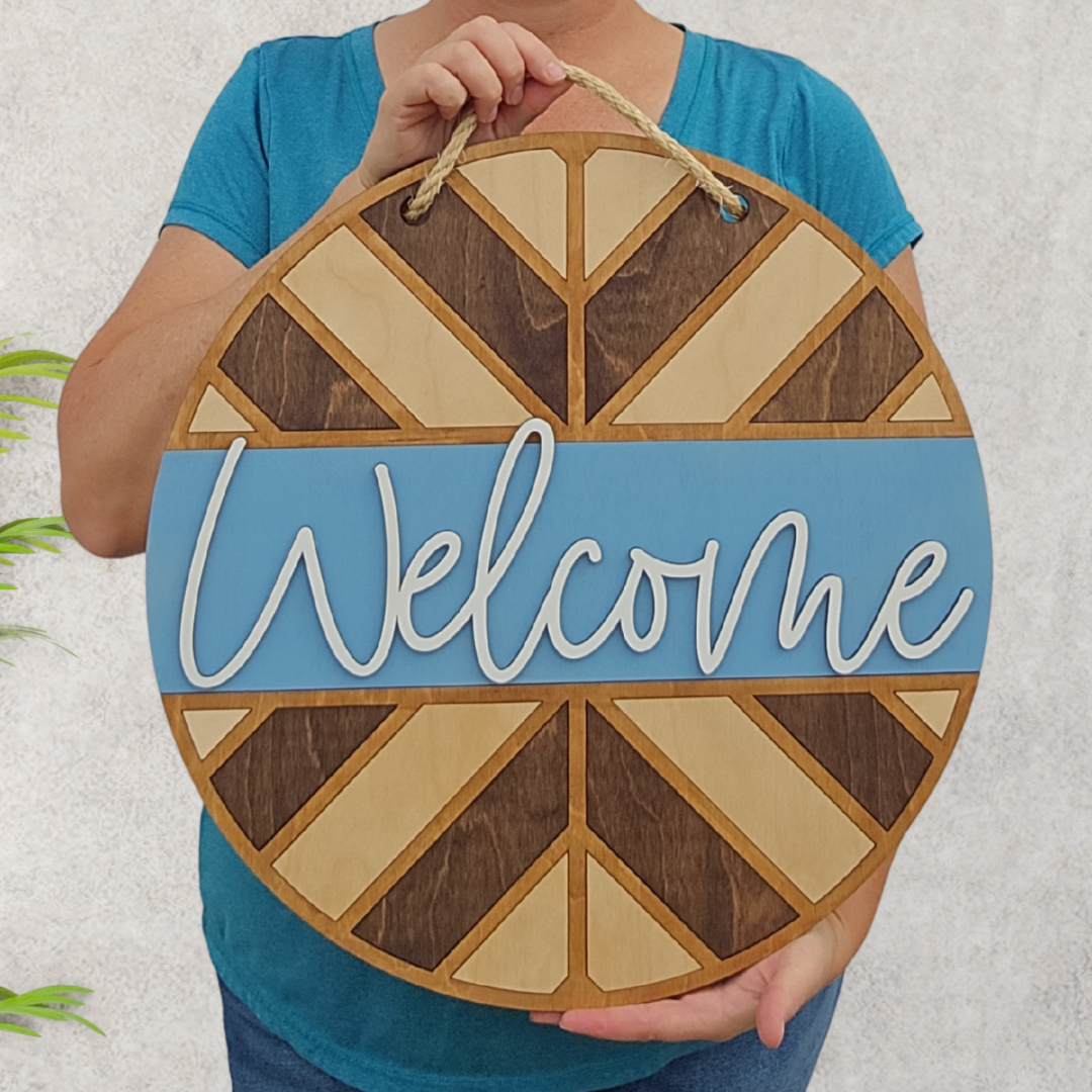 A person holds a round "Welcome" sign with geometric brown and tan patterns, blue background, and a rope handle.
