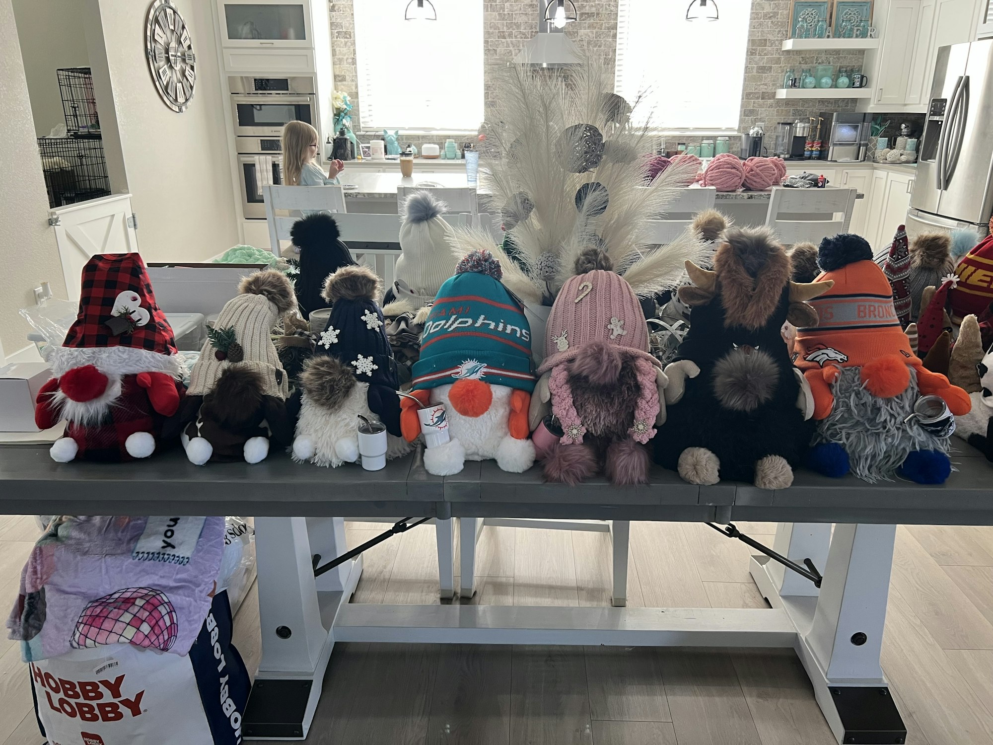 A row of decorative gnomes with various hats on a table, a pack of yarn in the background, and a child sitting in the kitchen.