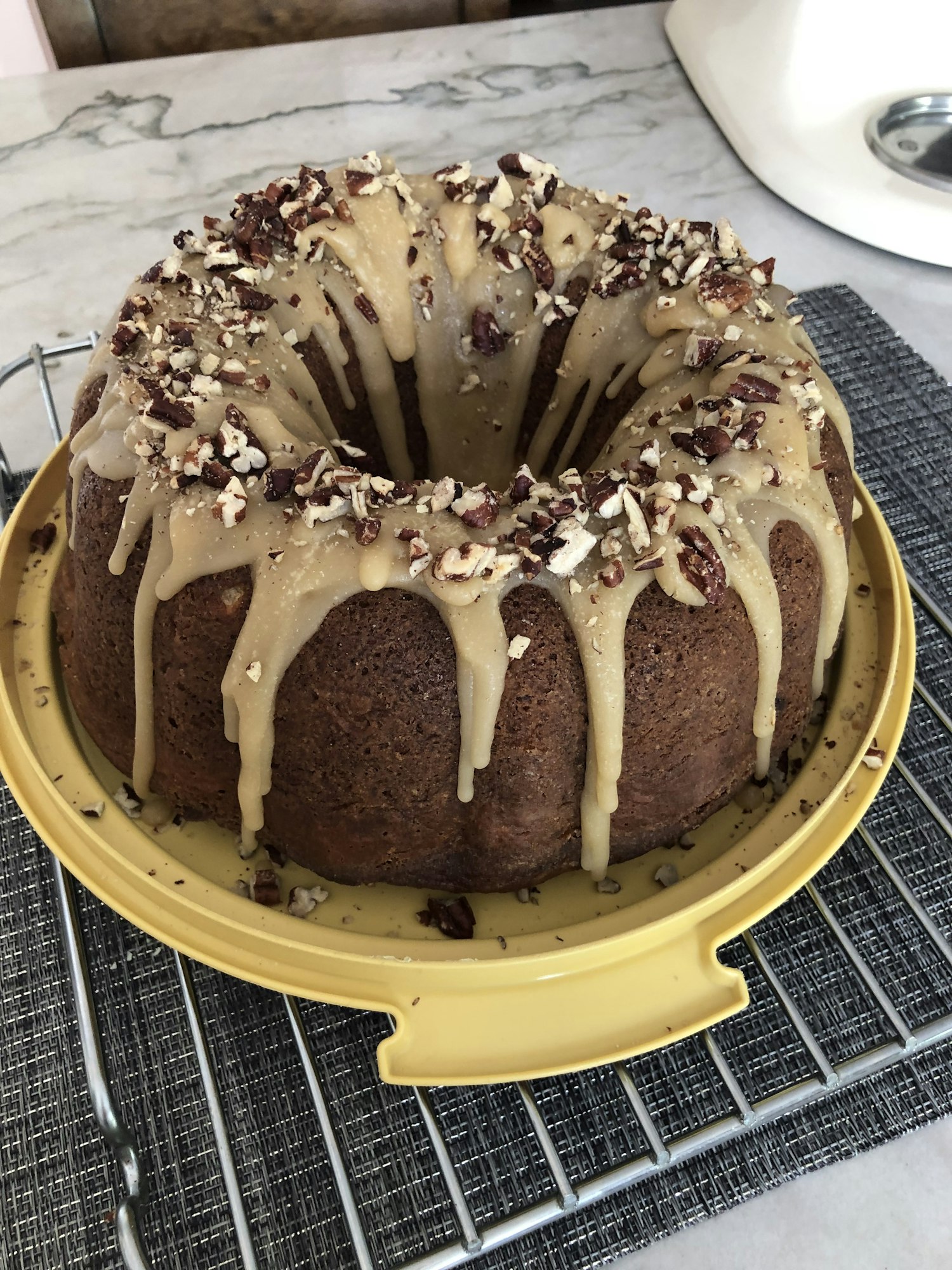 A bundt cake with glaze and chopped nuts on top, placed on a yellow platter and a cooling rack.
