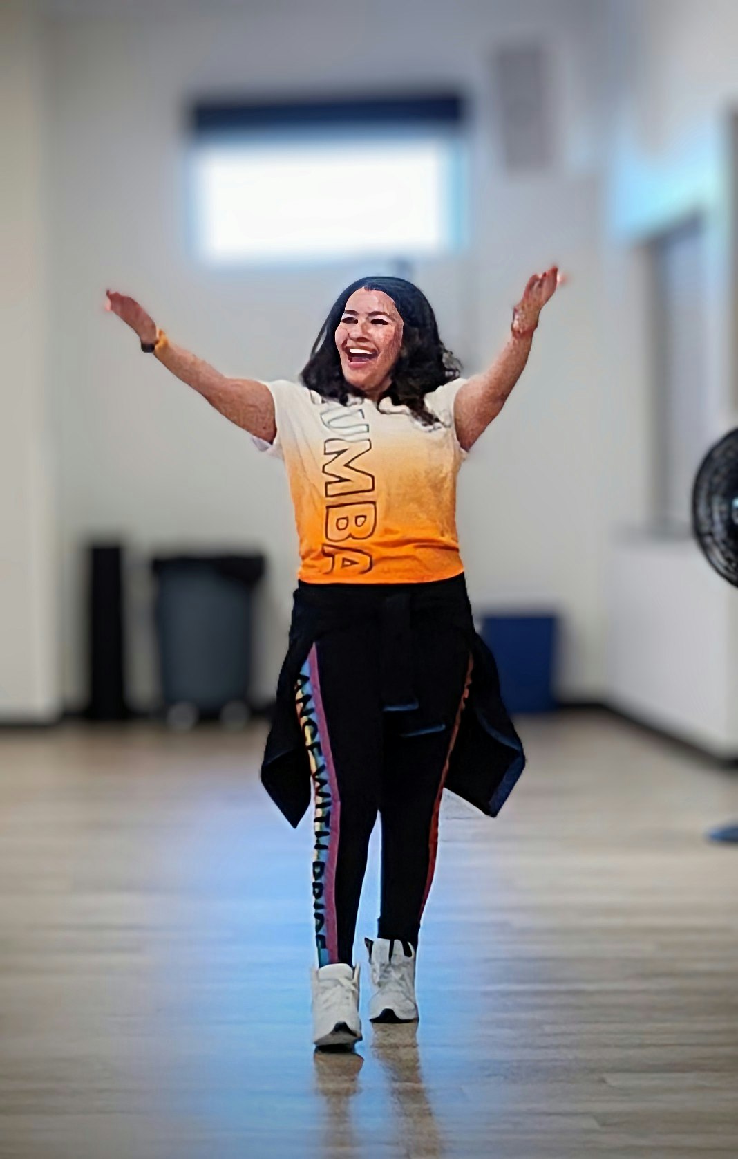 A person happily doing Zumba, arms raised, wearing a Zumba T-shirt in a brightly lit room.