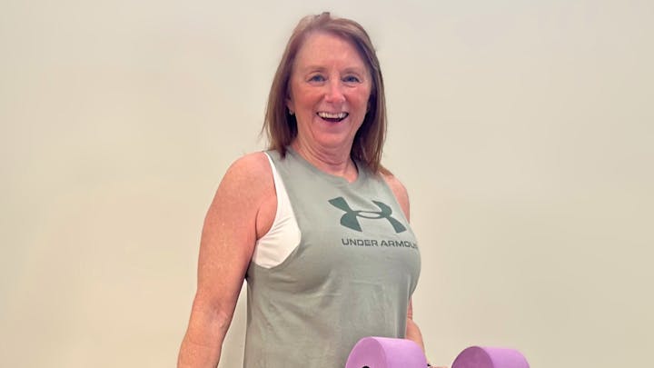 A person wearing an Under Armour tank top, smiling, holding a purple yoga mat.