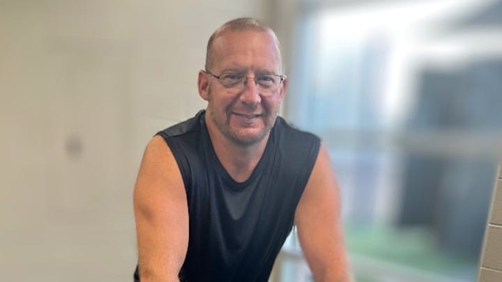 A man with short hair and glasses is smiling while wearing a sleeveless black shirt in a bright indoor setting.