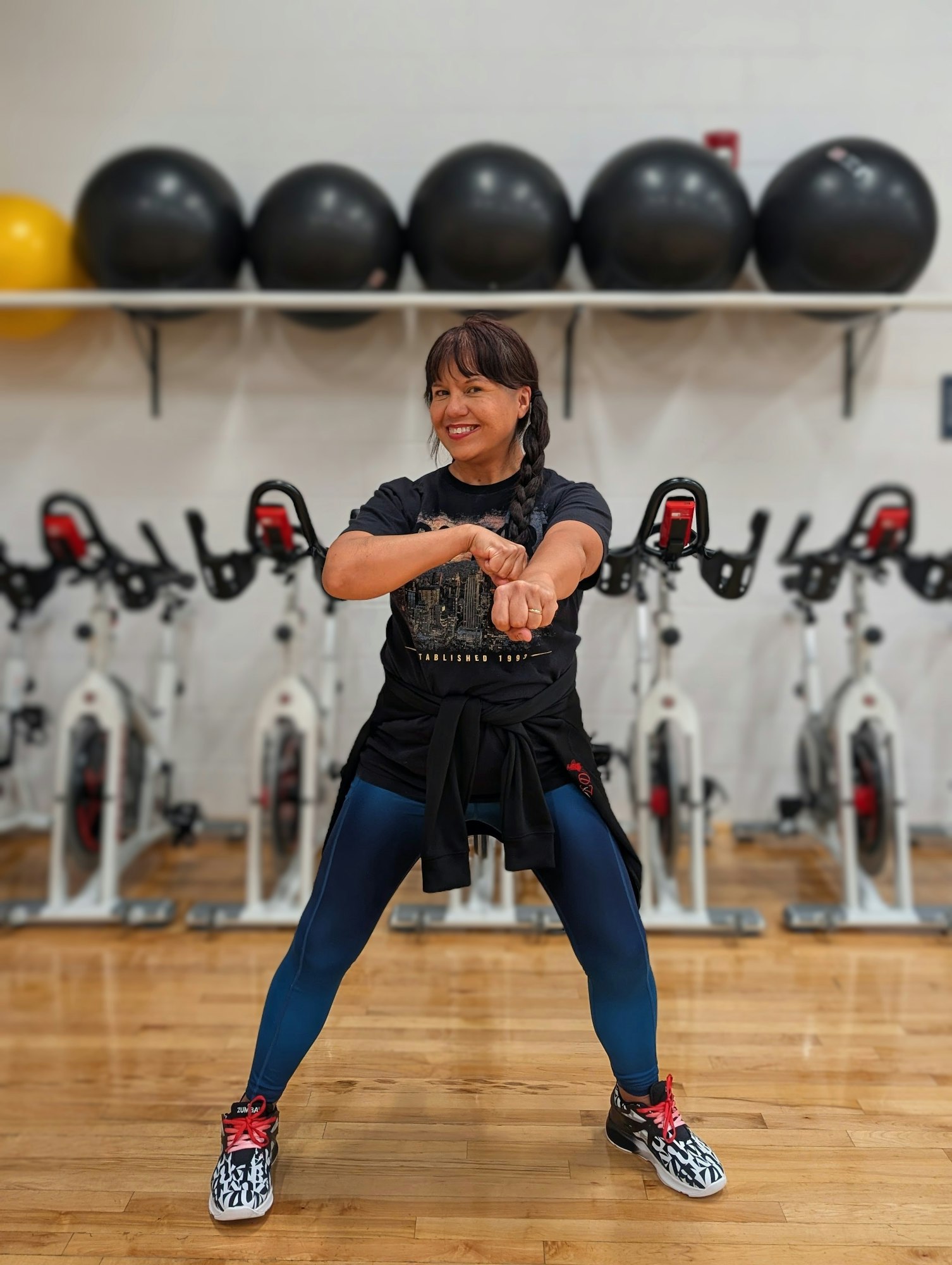 A person posing happily in a gym with exercise bikes, wearing workout attire and standing on a wooden floor.