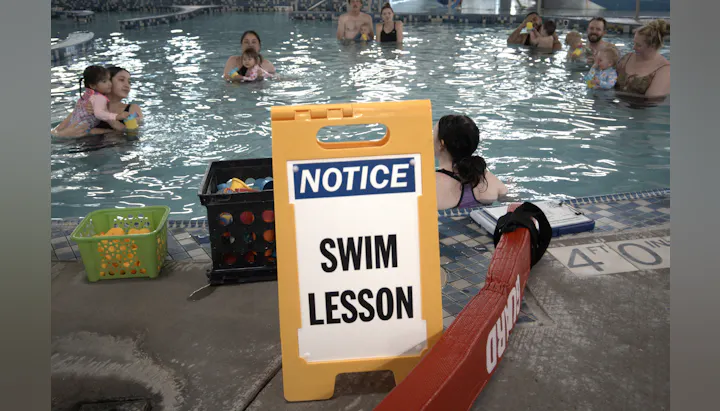 A swim lesson in progress at a pool, with participants and a notice sign. There are children and adults enjoying the activity.