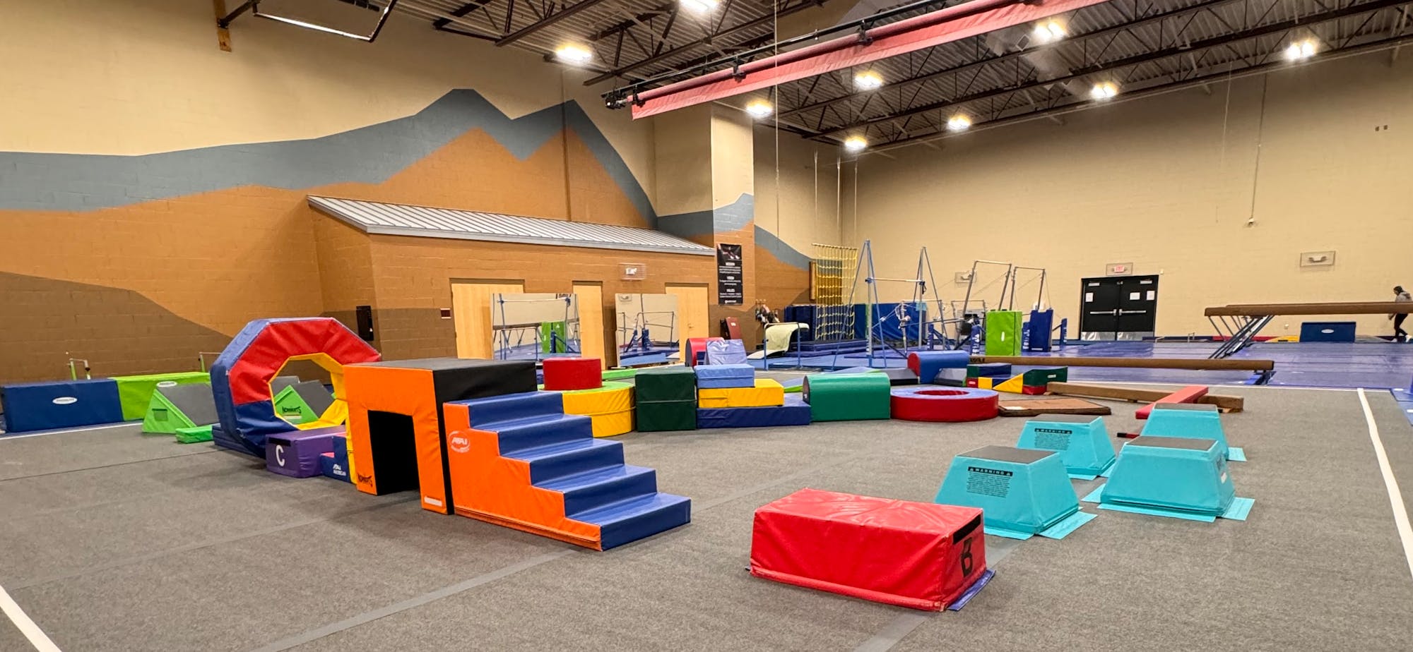 The image shows a colorful gymnasium layout with various foam blocks and equipment for gymnastics and obstacle courses.