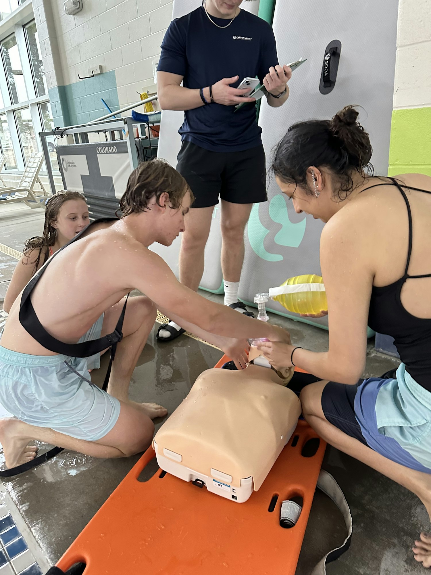 CPR training session with an instructor and participants practicing on a mannequin by the poolside.