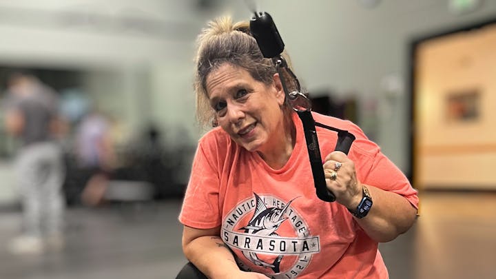 A person in a pink shirt at a gym, holding an exercise strap, smiling at the camera.