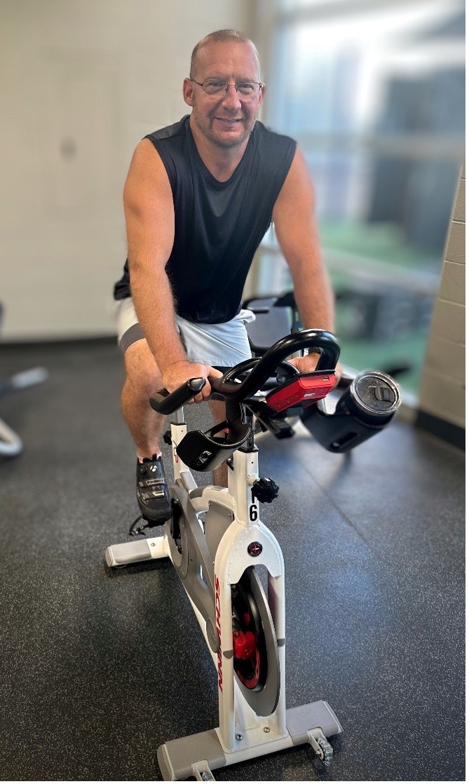 Person on an exercise bike in a gym, wearing a sleeveless shirt and glasses, smiling at the camera.