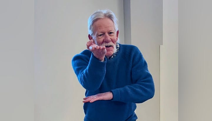 An older man in a blue sweater gestures with his hands, possibly explaining or performing something.
