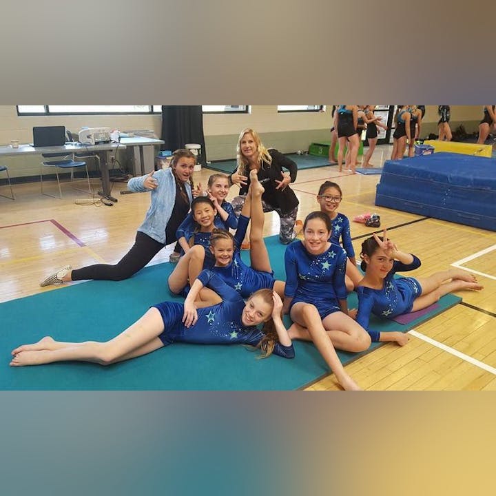 A group of young gymnasts in blue leotards poses playfully on a mat, with some stretching and others making fun gestures.
