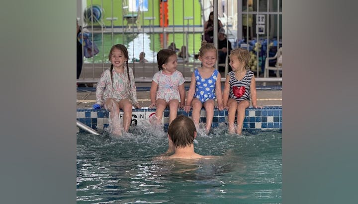 Four young girls in swimsuits sit on the edge of a pool, smiling and splashing water, while an adult is swimming nearby.