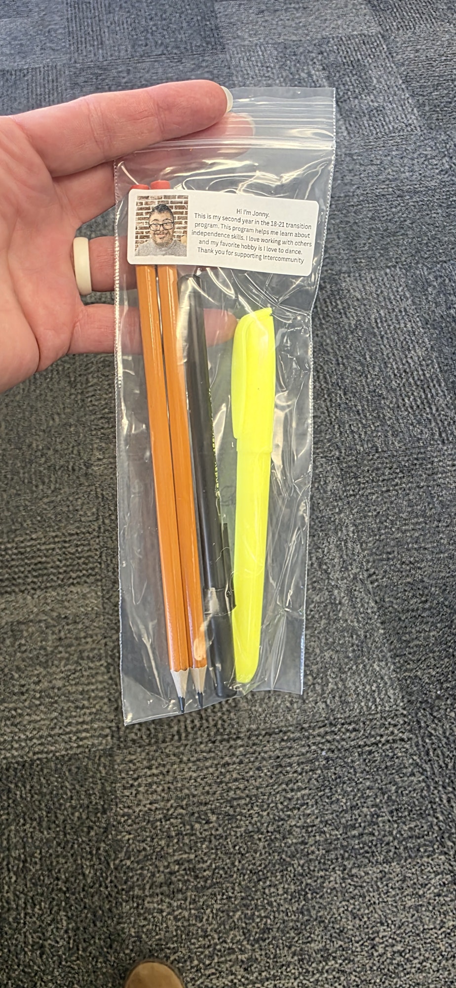 A hand holds a clear plastic bag containing pencils, a highlighter, and a note with a photo and text.