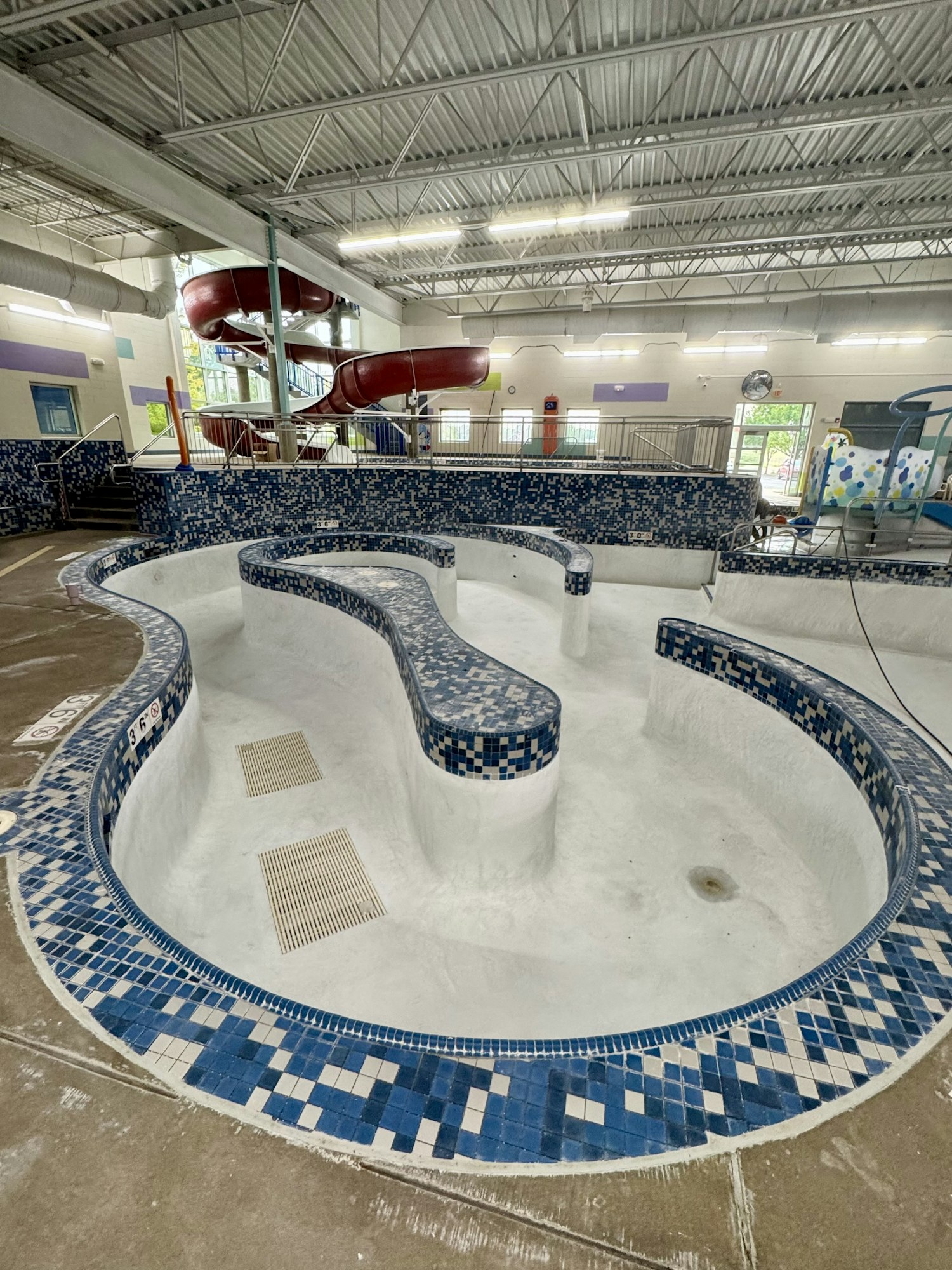 An empty indoor pool area with blue-tiled edges, a spiral red slide, and overhead metal beams.