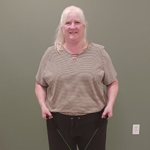 A person with long hair is smiling and holding out the waistband of their pants. They are standing against a plain wall.