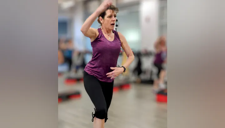 A woman in a purple tank top exercises energetically in a gym, with others engaged in fitness activities in the background.