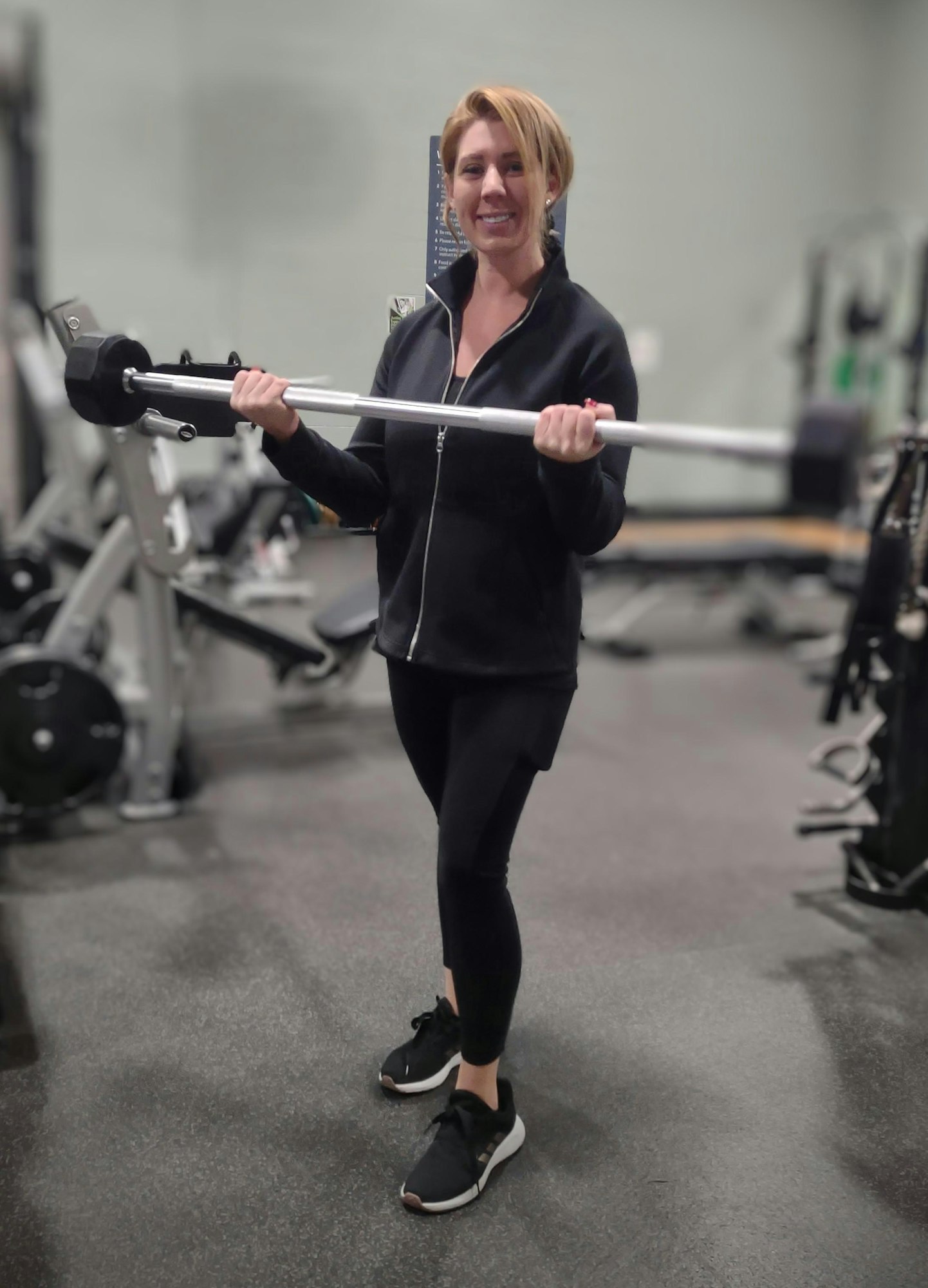 A smiling woman holding a barbell in a gym setting.