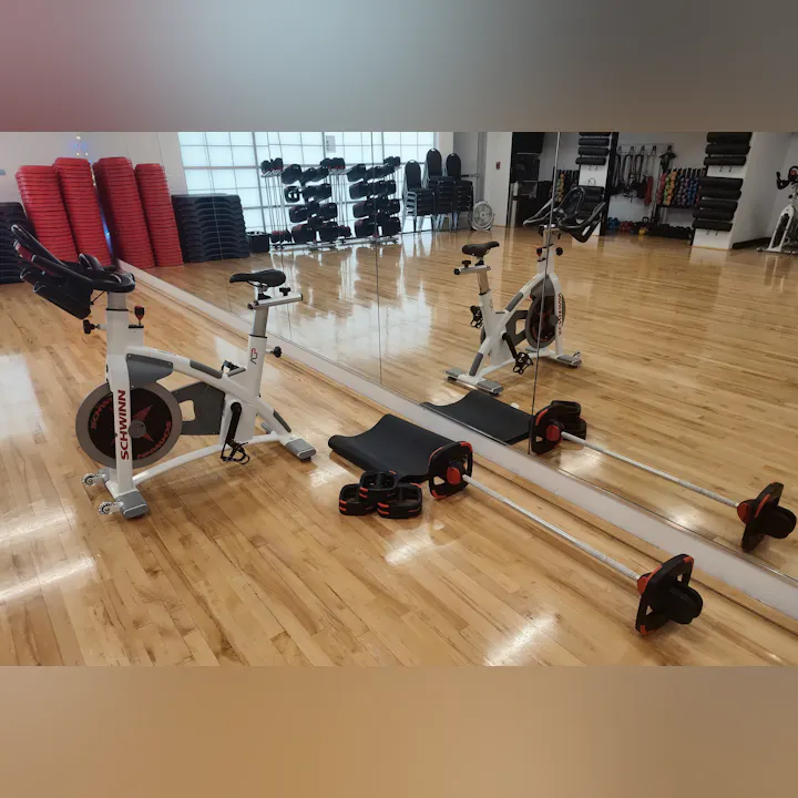 Image of a fitness studio featuring a Schwinn spinning bike, weights, a mat, and mirrored walls, with a wooden floor.