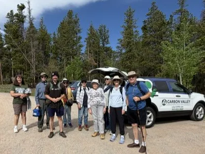 A group of hikers poses together outdoors near trees and a vehicle, ready for an adventure in a natural setting.