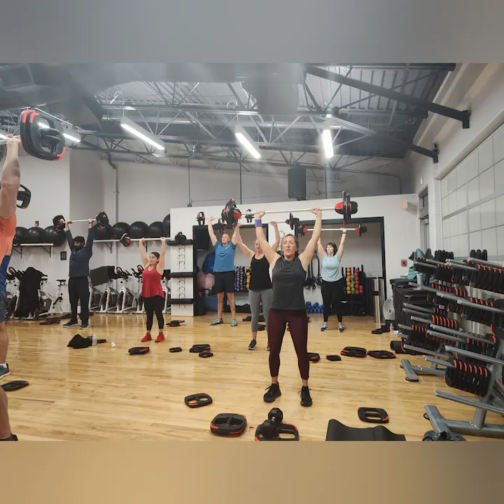 A group workout session in a gym, participants are engaged in lifting weights overhead on a wooden floor.