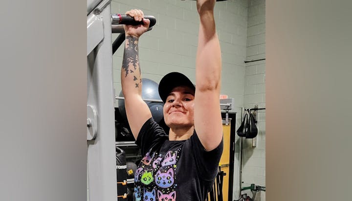 A person is doing pull-ups at a gym, smiling while wearing a playful cat-themed shirt and a cap. Fitness equipment is in the background.