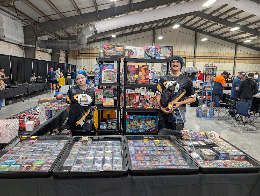 Two people at a card game booth selling trading cards and boxed games in a convention hall.