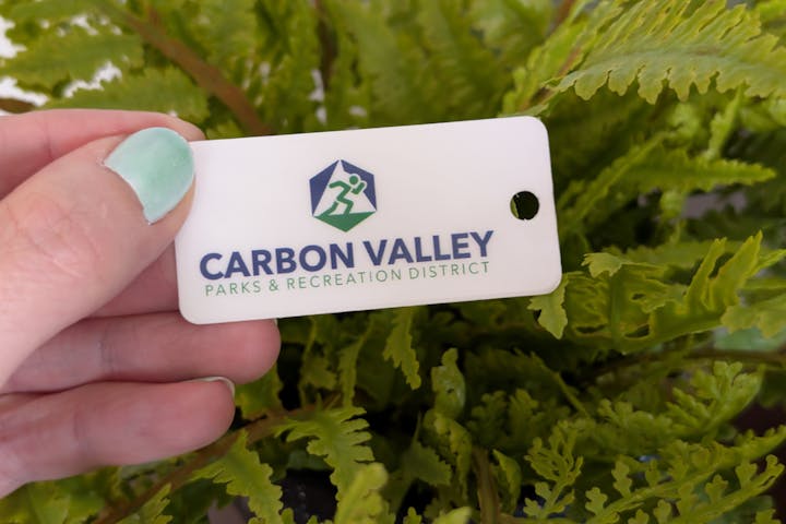 A hand holding a membership card for Carbon Valley Parks & Recreation District, with green ferns in the background.