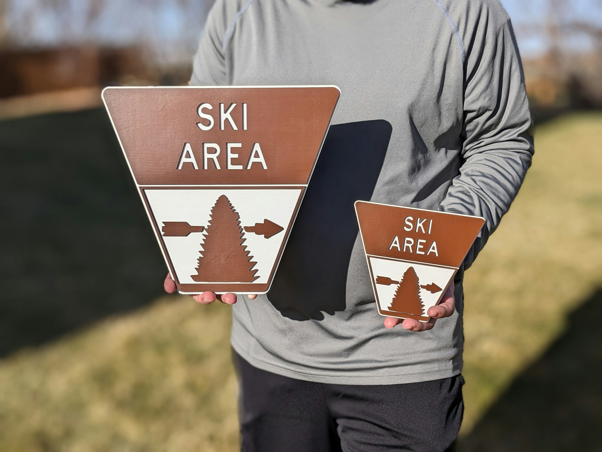 Person holding two triangular "Ski Area" signs, one large and one small, with tree and arrows symbols.