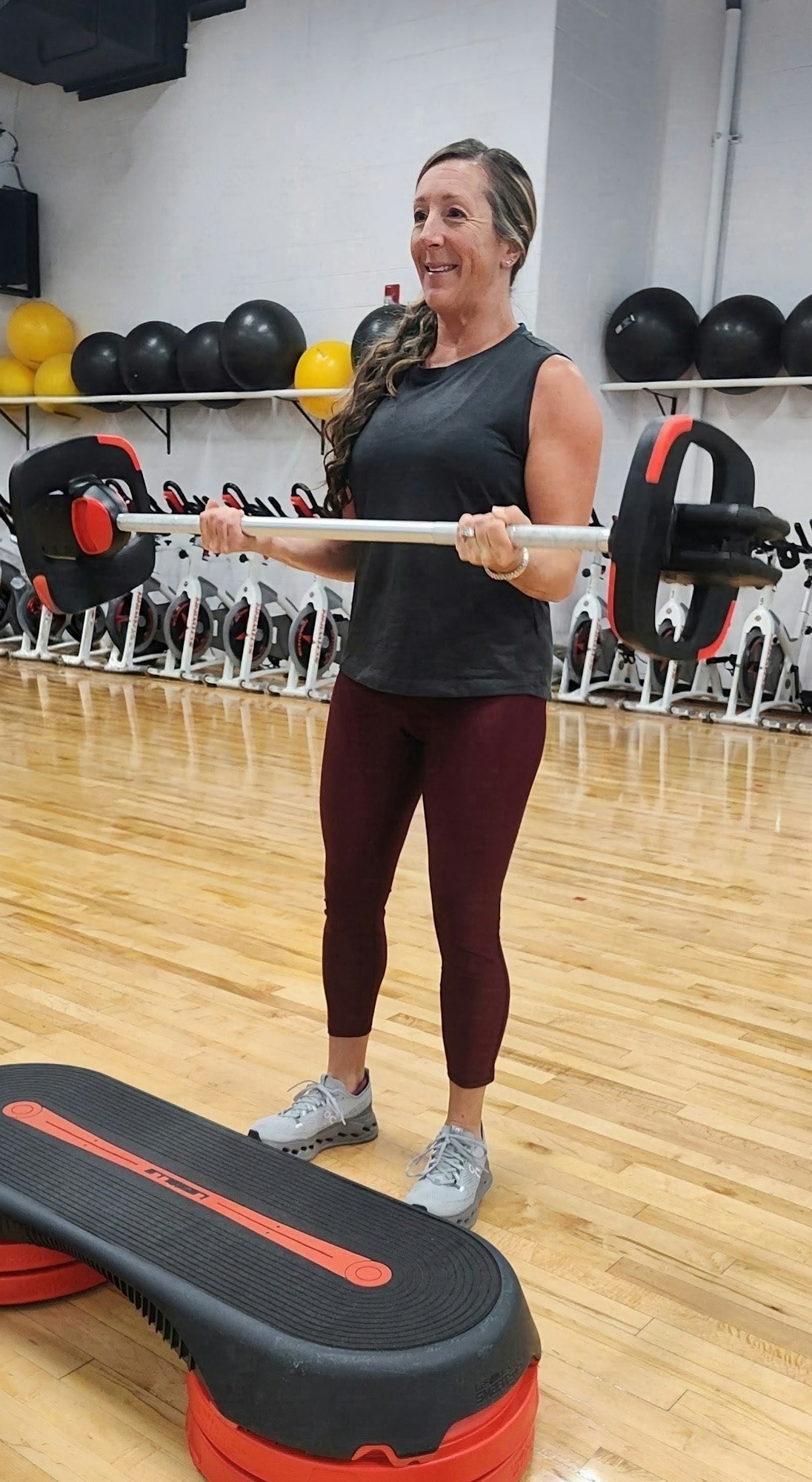 A person is smiling while lifting a barbell in a gym, standing over a step platform with exercise equipment in the background.