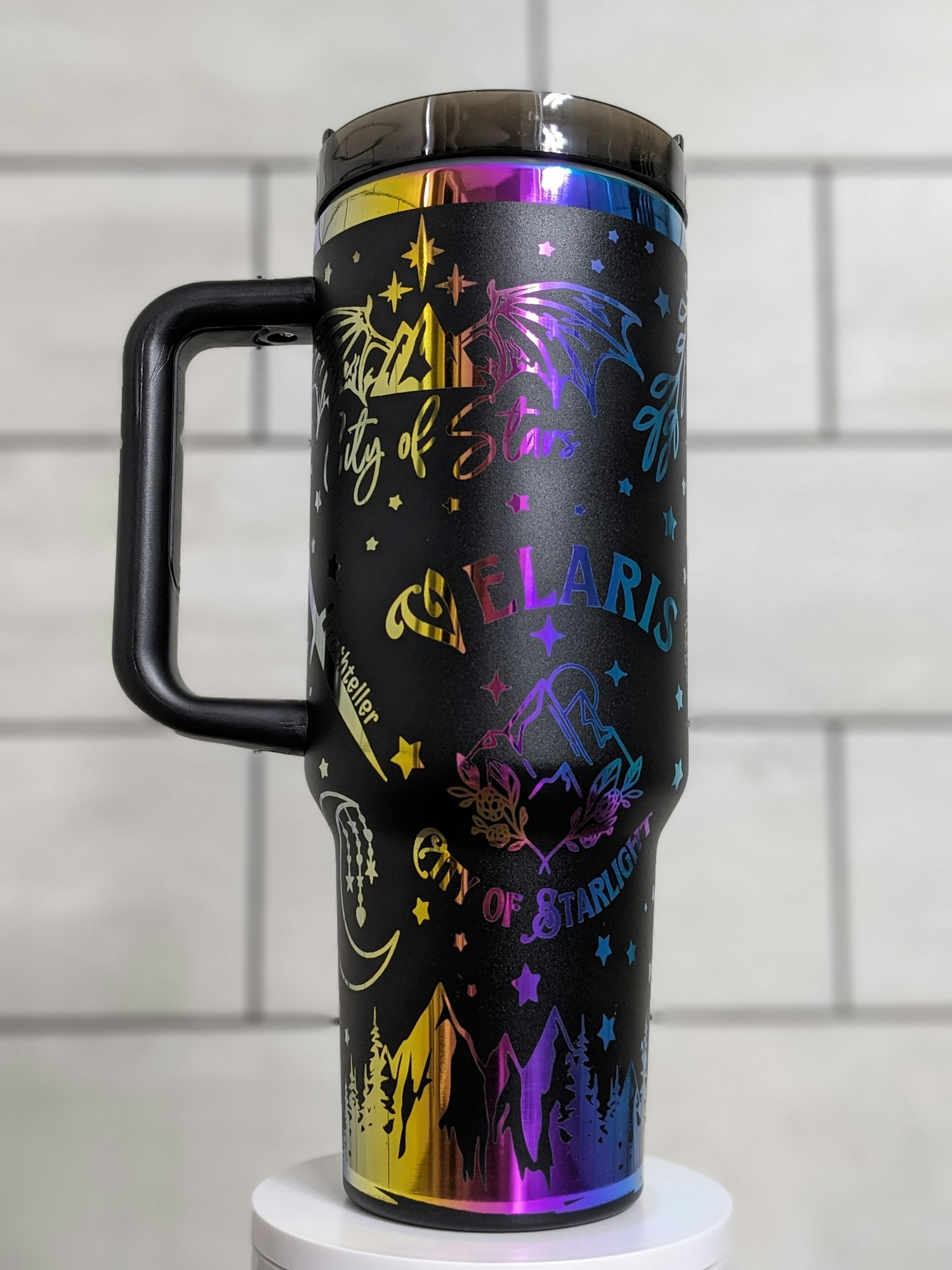 A black travel mug with colorful designs, including stars, mountains, a dragon, and text like "City of Stars" and "VELARIS."
