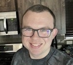 A person with glasses smiling in a kitchen, with modern appliances and wooden cabinetry in the background.