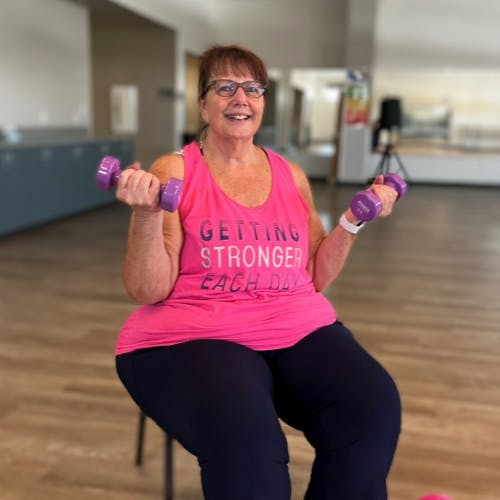 A woman in a gym setting is sitting on a chair, smiling and holding purple dumbbells. Her pink tank top reads "Getting Stronger Each Day."