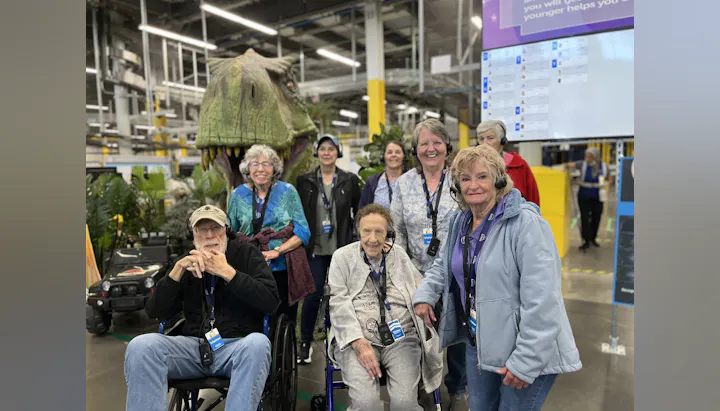 A group of elderly visitors, some in wheelchairs, pose in front of a dinosaur exhibit in a lively indoor setting.
