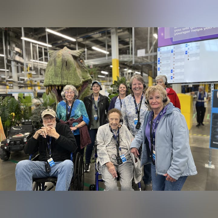 A group of elderly visitors, some in wheelchairs, pose in front of a dinosaur exhibit in a lively indoor setting.
