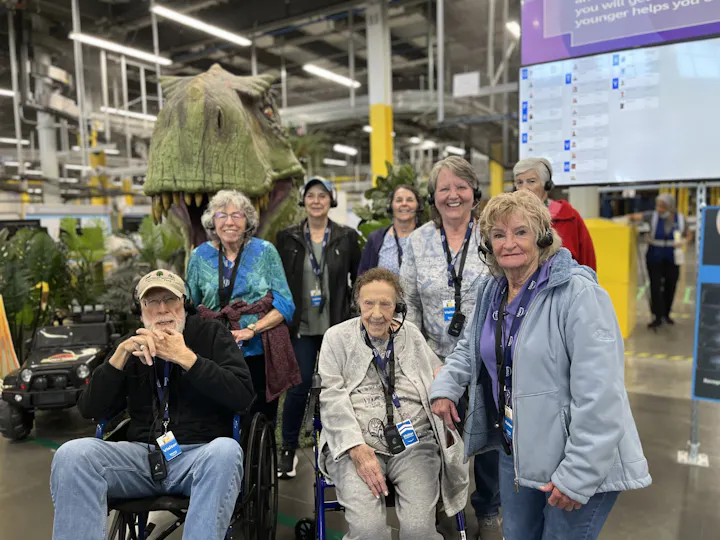 A group of elderly visitors, some in wheelchairs, pose in front of a dinosaur exhibit in a lively indoor setting.
