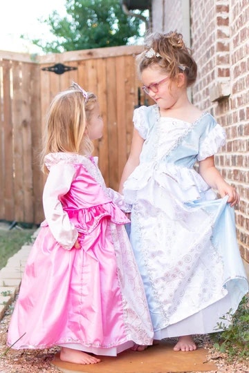 Two young girls dressed as princesses, one in a pink dress and the other in blue, look at each other outside by a wooden fence.