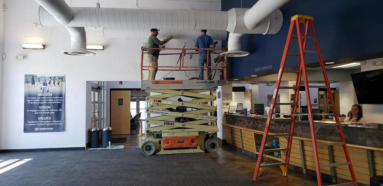 Two workers are on a scissor lift, painting the ceiling in a lobby area with a red ladder and a service desk visible.